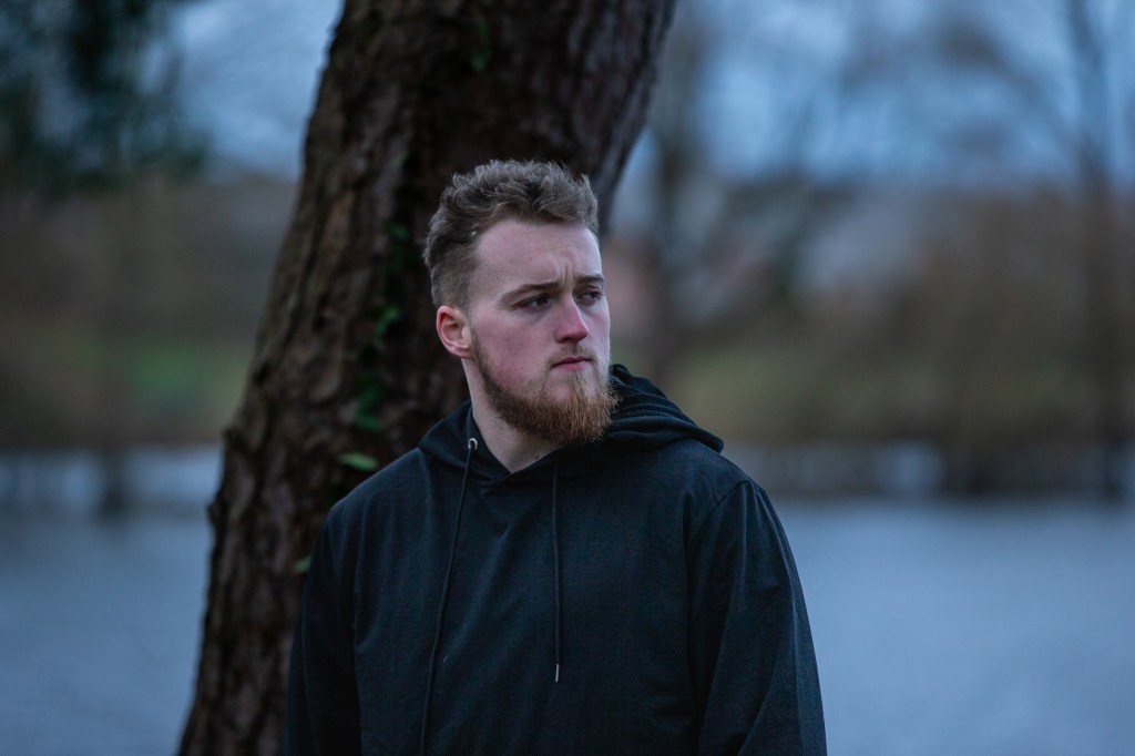 A man wearing a black hoodie with a beard and a serious expression standing beside a tree looking into the distance