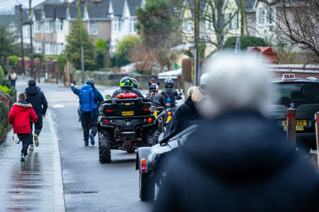 Image showing a cavalcade of motorcycles and quad bikes following a man walking with a blue rucksack on his back and his hand raised videoing the people behind him